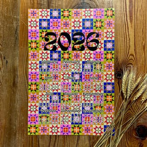 Puede incluir: Un colorido calendario de 2026 con un patrón geométrico en rosa, azul, naranja y verde. El calendario muestra los meses del año y está sobre una superficie de madera. Un manojo de espigas de trigo descansa a la derecha.