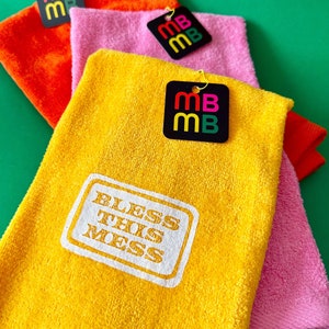 May include: Three yellow, pink, and orange towels with the text "Bless This Mess" printed on the yellow towel. Each towel has a black tag with the letters "MB" in red, green, and yellow.