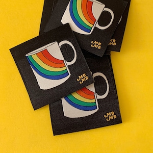 May include: A set of four black fabric patches featuring a rainbow mug design with the text "JMB JMB" in gold embroidery.