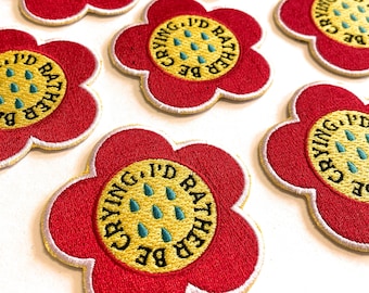 May include: Six red flower-shaped iron-on patches with white outlines. Each patch has a yellow circle in the center with the text "BE CRYING. I'D RATHER" in a black outline. The yellow circle also has five green teardrop shapes.