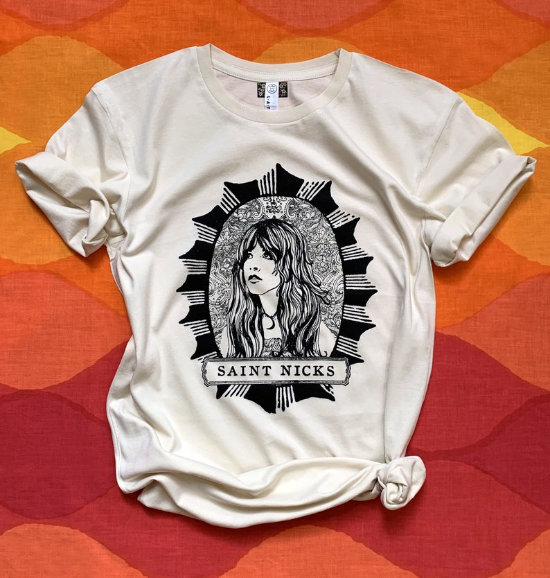 K&ouml;nnte beinhalten: Ein cremefarbenes T-Shirt mit einer schwarz-wei&szlig;en Illustration einer Frau mit langen Haaren in einem Kreis mit einem Sternenausbruch-Design. Der Text "SAINT NICKS" ist unter der Illustration gedruckt.