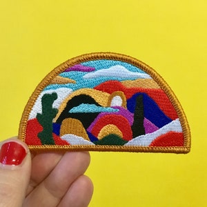 May include: A colourful embroidered patch depicting a desert landscape with mountains, a cactus, and a rainbow. The patch is shaped like a half-circle and has a gold border.