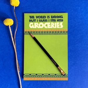 May include: A rectangular green notepad featuring the text THE WORLD IS ENDING BUT I GUESS I STILL NEED GROCERIES in dark green and white. The notepad has decorative borders and a black pencil with a gold ferrule resting diagonally across it.