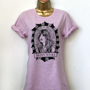K&ouml;nnte beinhalten: Ein helllila T-Shirt mit einer schwarz-wei&szlig;en Illustration einer Frau mit langen Haaren. Die Illustration ist von einem schwarzen und wei&szlig;en kreisf&ouml;rmigen Design umgeben, mit dem Text "SAINT NICKS" am unteren Rand.