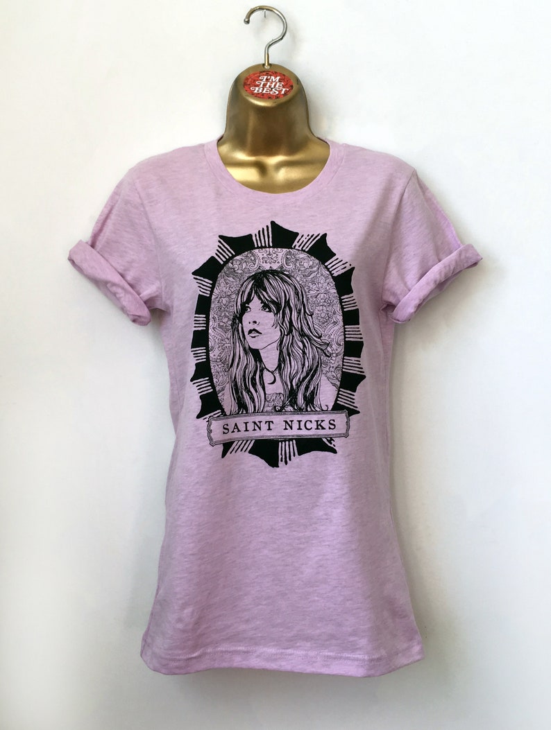 K&ouml;nnte beinhalten: Ein helllila T-Shirt mit einer schwarz-wei&szlig;en Illustration einer Frau mit langen Haaren. Die Illustration ist von einem schwarzen und wei&szlig;en kreisf&ouml;rmigen Design umgeben, mit dem Text "SAINT NICKS" am unteren Rand.