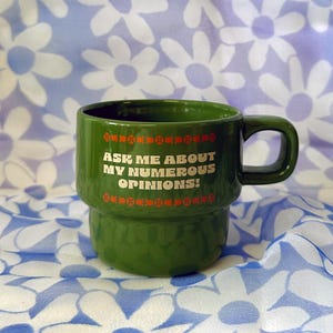 Peut inclure: Une tasse en céramique verte avec une inscription blanche qui dit "Ask me about my numerous opinions!" La tasse a une bordure décorative blanche et orange.