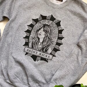 May include: Grey sweatshirt with a black and white illustration of a woman with long hair in a circular frame with the text "SAINT NICKS" at the bottom.