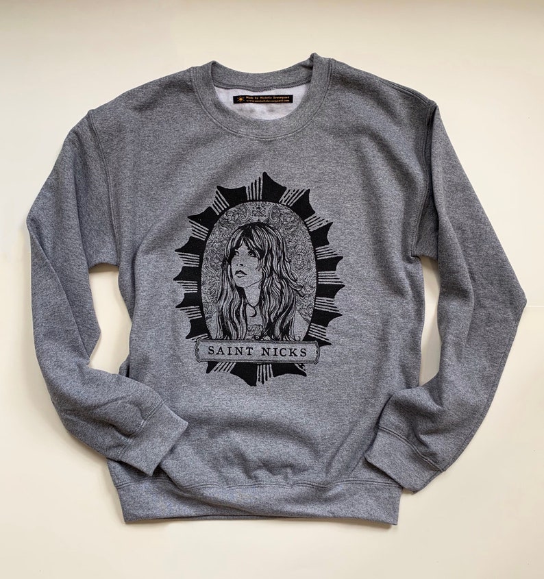 May include: Grey sweatshirt featuring a black and white illustration of a woman with long hair in a circle with a starburst design. The text "SAINT NICKS" is printed below the illustration.