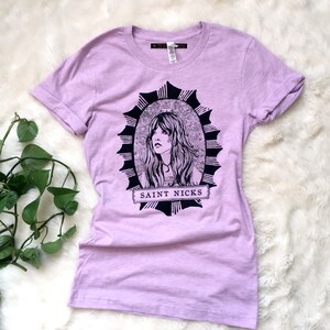 K&ouml;nnte beinhalten: Ein lavendelfarbenes T-Shirt mit einer schwarz-wei&szlig;en Illustration einer Frau mit langen Haaren in einem runden Rahmen. Der Text "SAINT NICKS" ist unter dem Rahmen gedruckt.