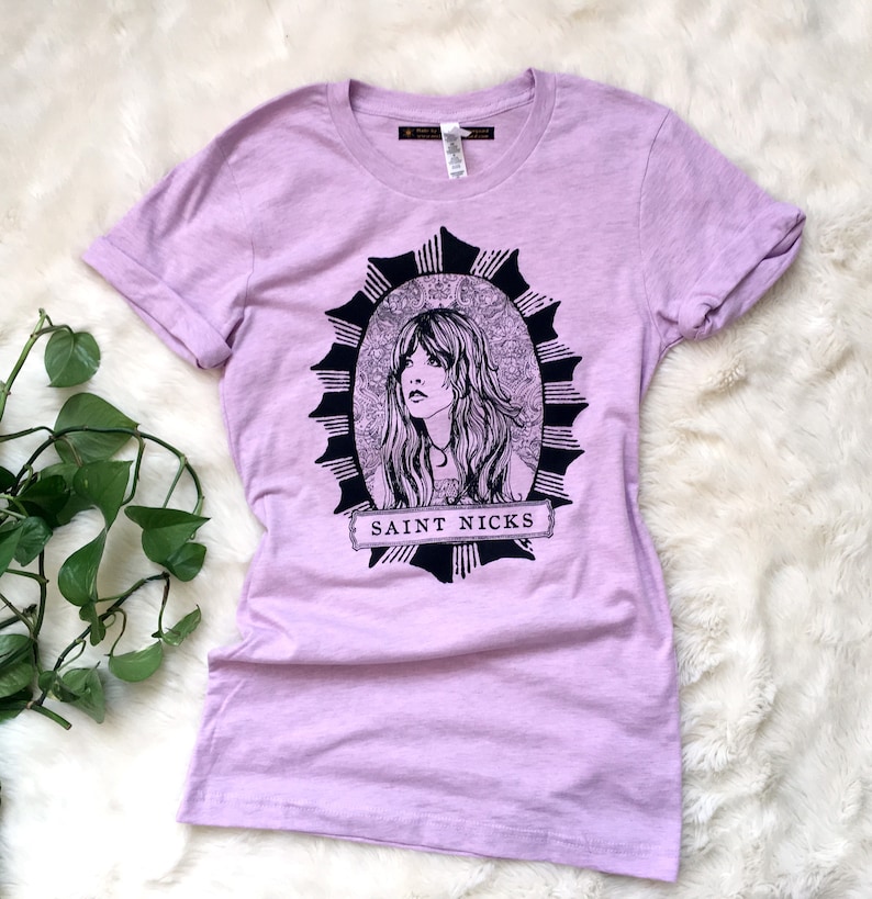 K&ouml;nnte beinhalten: Ein lavendelfarbenes T-Shirt mit einer schwarz-wei&szlig;en Illustration einer Frau mit langen Haaren in einem runden Rahmen. Der Text "SAINT NICKS" ist unter dem Rahmen gedruckt.