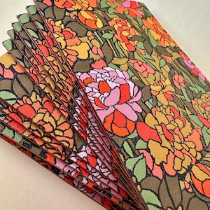 May include: A stack of 10 fabric coasters with a floral pattern in shades of orange, pink, yellow, and green on a brown background.