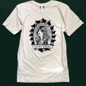 K&ouml;nnte beinhalten: Ein cremefarbenes T-Shirt mit einer schwarz-wei&szlig;en Illustration einer Frau mit langen Haaren in einem Kreis mit einem Sternenausbruch-Design. Der Text "SAINT NICKS" ist unter der Illustration gedruckt.