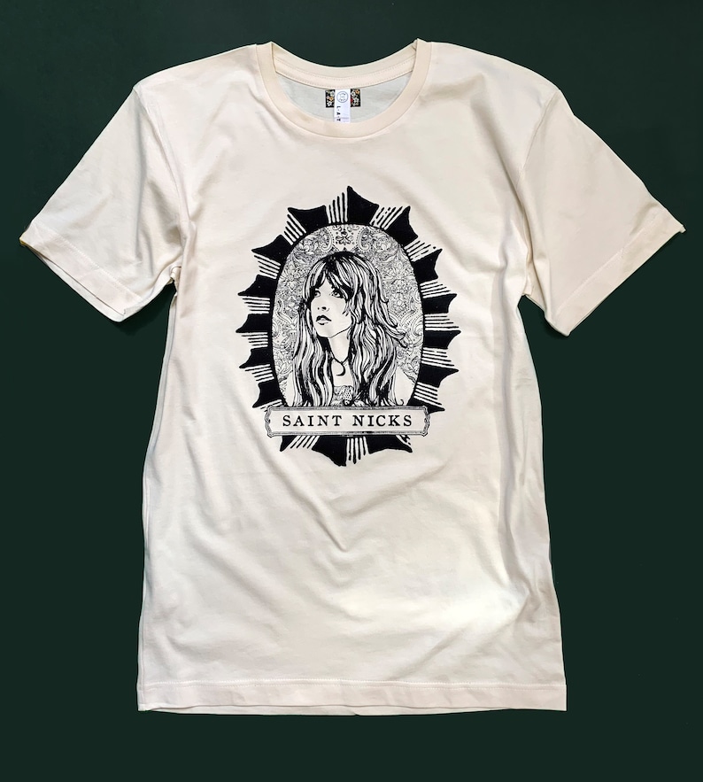 K&ouml;nnte beinhalten: Ein cremefarbenes T-Shirt mit einer schwarz-wei&szlig;en Illustration einer Frau mit langen Haaren in einem Kreis mit einem Sternenausbruch-Design. Der Text "SAINT NICKS" ist unter der Illustration gedruckt.