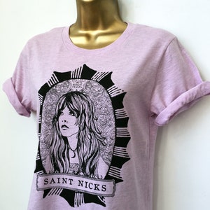 K&ouml;nnte beinhalten: Ein helles lila T-Shirt mit einer schwarz-wei&szlig;en Illustration einer Frau mit langen Haaren. Die Illustration ist von einem schwarzen und wei&szlig;en, verzierten Rahmen umgeben. Der Text "SAINT NICKS" ist unter der Illustration gedruckt.
