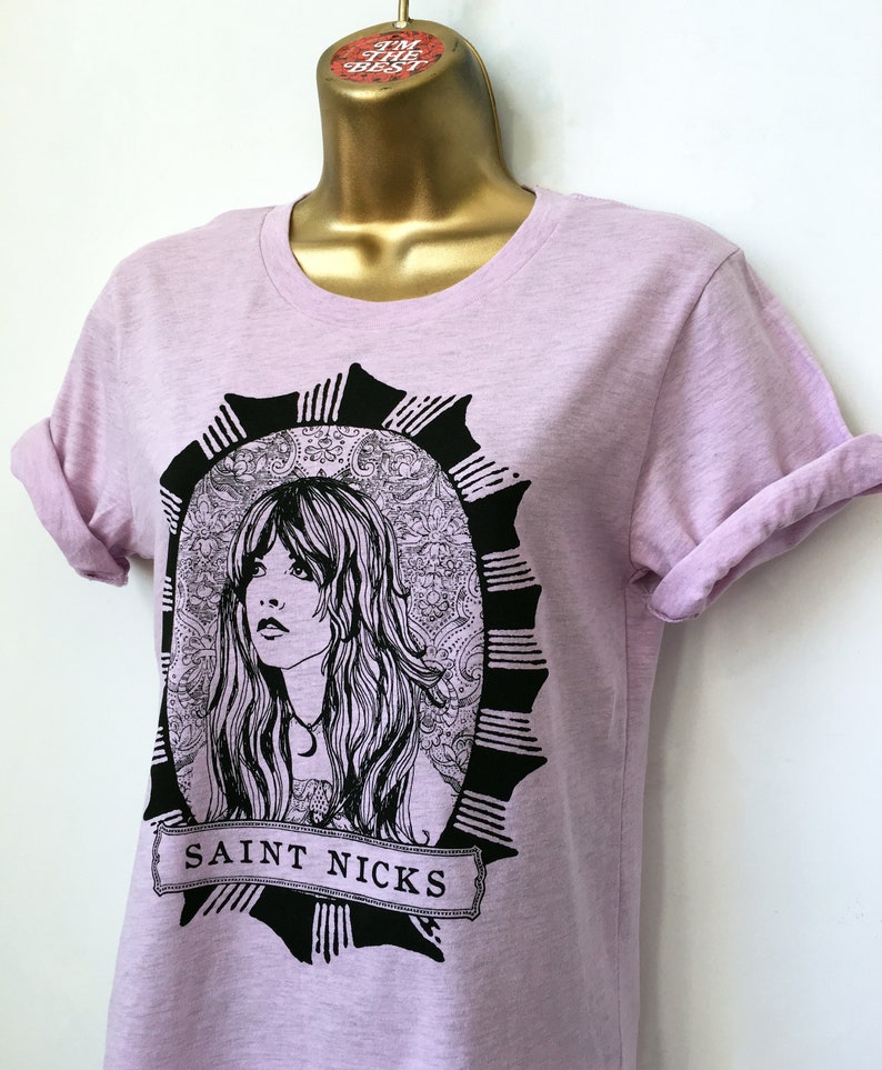 K&ouml;nnte beinhalten: Ein helles lila T-Shirt mit einer schwarz-wei&szlig;en Illustration einer Frau mit langen Haaren. Die Illustration ist von einem schwarzen und wei&szlig;en, verzierten Rahmen umgeben. Der Text "SAINT NICKS" ist unter der Illustration gedruckt.
