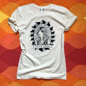 K&ouml;nnte beinhalten: Ein wei&szlig;es T-Shirt mit einer schwarz-wei&szlig;en Illustration einer Frau mit langen Haaren in einem Kreis mit einem Sternenausbruch-Design. Der Text "SAINT NICKS" ist unter der Illustration gedruckt.