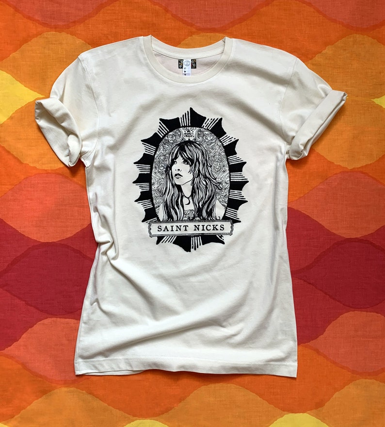 K&ouml;nnte beinhalten: Ein wei&szlig;es T-Shirt mit einer schwarz-wei&szlig;en Illustration einer Frau mit langen Haaren in einem Kreis mit einem Sternenausbruch-Design. Der Text "SAINT NICKS" ist unter der Illustration gedruckt.