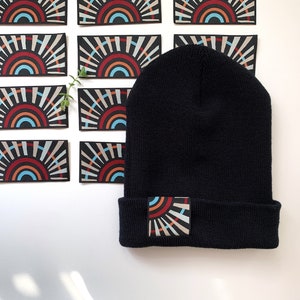 Puede incluir: Gorro negro con borde doblado, con un parche rectangular con un diseño de sol colorido. El diseño incluye rayas rojas, naranjas, azules y blancas. Varios parches similares están dispuestos alrededor del gorro.