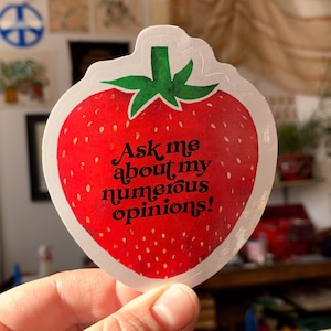 May include: A red strawberry sticker with a white outline. The sticker has the text "Ask me about my numerous opinions!" in black.
