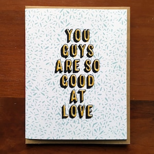 May include: A white card with a blue and white patterned background. The card has black text that reads "YOU GUYS ARE SO GOOD AT LOVE" with the words "GOOD" and "LOVE" in yellow.