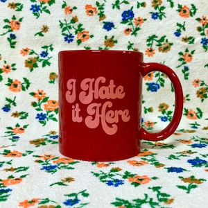 May include: Red ceramic mug with a white floral pattern background. The mug has a white handle and says "I Hate it Here" in pink cursive lettering.