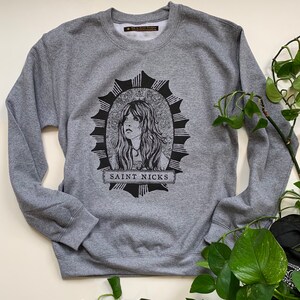May include: Grey sweatshirt with a black and white graphic print of a woman's face in a circle with the text "SAINT NICKS" below.