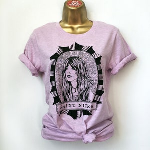 K&ouml;nnte beinhalten: Ein lavendelfarbenes T-Shirt mit einer Schwarz-Wei&szlig;-Illustration einer Frau mit langen Haaren. Die Illustration ist von einem dekorativen Rahmen umgeben und der Text "SAINT NICKS" befindet sich unter dem Bild.