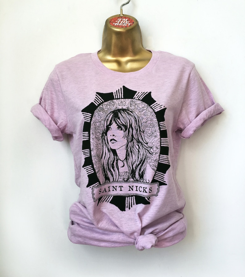 K&ouml;nnte beinhalten: Ein lavendelfarbenes T-Shirt mit einer Schwarz-Wei&szlig;-Illustration einer Frau mit langen Haaren. Die Illustration ist von einem dekorativen Rahmen umgeben und der Text "SAINT NICKS" befindet sich unter dem Bild.
