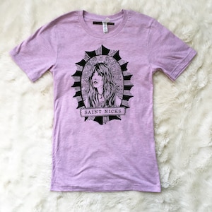 K&ouml;nnte beinhalten: Ein lavendelfarbenes T-Shirt mit einer schwarz-wei&szlig;en Illustration einer Frau mit langen Haaren in einem runden Rahmen. Der Text "SAINT NICKS" ist auf einem Banner unter dem Rahmen gedruckt.