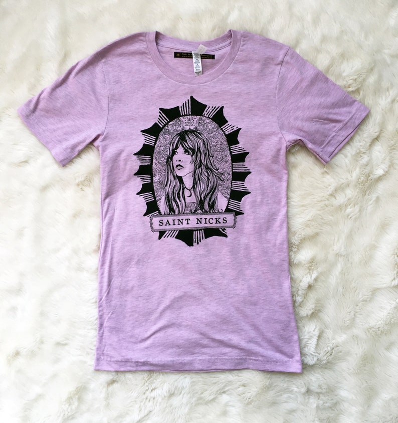 K&ouml;nnte beinhalten: Ein lavendelfarbenes T-Shirt mit einer schwarz-wei&szlig;en Illustration einer Frau mit langen Haaren in einem runden Rahmen. Der Text "SAINT NICKS" ist auf einem Banner unter dem Rahmen gedruckt.