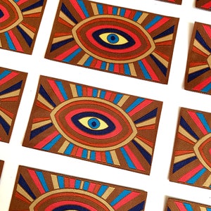 Könnte beinhalten: Eine Sammlung bunter Augen-Patches mit geometrischem Design. Die Patches zeigen ein großes Auge mit konzentrischen Kreisen in Rot-, Blau-, Gelb- und Brauntönen. Der Hintergrund ist ein brauner Stoff mit einem strahlenförmigen Muster.