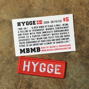 May include: Red iron-on patch with white text that reads "HYGGE". The patch is on a brown surface. The text on the card reads "HYGGE [HOO-GA'] "A DEEP SENSE OF PLACE & WELL-BEING; A FEELING OF FRIENDSHIP, WARMTH, CONTENTMENT AND PEACE WITH YOUR IMMEDIATE SURROUNDINGS. HYGGE IS A DANISH CONCEPT WHICH ROUGHLY DESCRIBES THAT WARM AND FUZZY FEELING WHEN YOU'RE SURROUNDED BY GOOD FOOD AND GOOD COMPANY. HYGGE IS AN IMPORTANT ELEMENT OF DANISH LIFE." MBMB MADE BY MICHELLE BRUSEGAARD WWW.MICHELLEBRUSEGAARD.COM"
