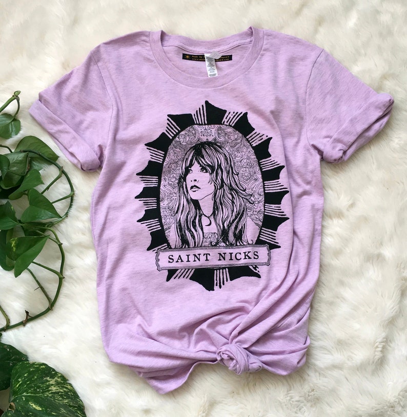 K&ouml;nnte beinhalten: Ein lavendelfarbenes T-Shirt mit einer schwarz-wei&szlig;en Illustration einer Frau mit langen Haaren. Die Illustration ist von einem schwarz-wei&szlig;en Sonnenstrahlen-Design umgeben. Der Text "SAINT NICKS" ist in Schwarz auf einem wei&szlig;en Banner unter der Illustration gedruckt.