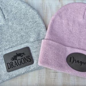May include: Two knit beanies with leather patches. One beanie is gray with a black dragon and the text "DRAGONS". The other beanie is pink with the text "Dragons".
