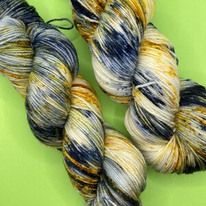 Può includere: Due gomitoli di filato in una combinazione di colori blu, bianco e giallo. Il filato è tinto a mano e ha un effetto maculato.