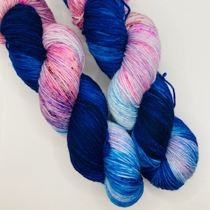 May include: Two skeins of yarn in a blue, pink, and white colorway. The yarn is hand-dyed and has a speckled appearance.