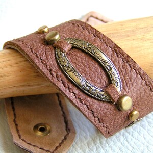 May include: A brown leather bracelet with a decorative oval metal clasp. The clasp has a floral design and is accented with small metal studs.