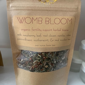 May include: A brown paper bag with a clear window displaying loose leaf tea. The label reads "WOMB BLOOM" and lists ingredients like raspberry leaf and red clover. The bag contains one ounce of tea.