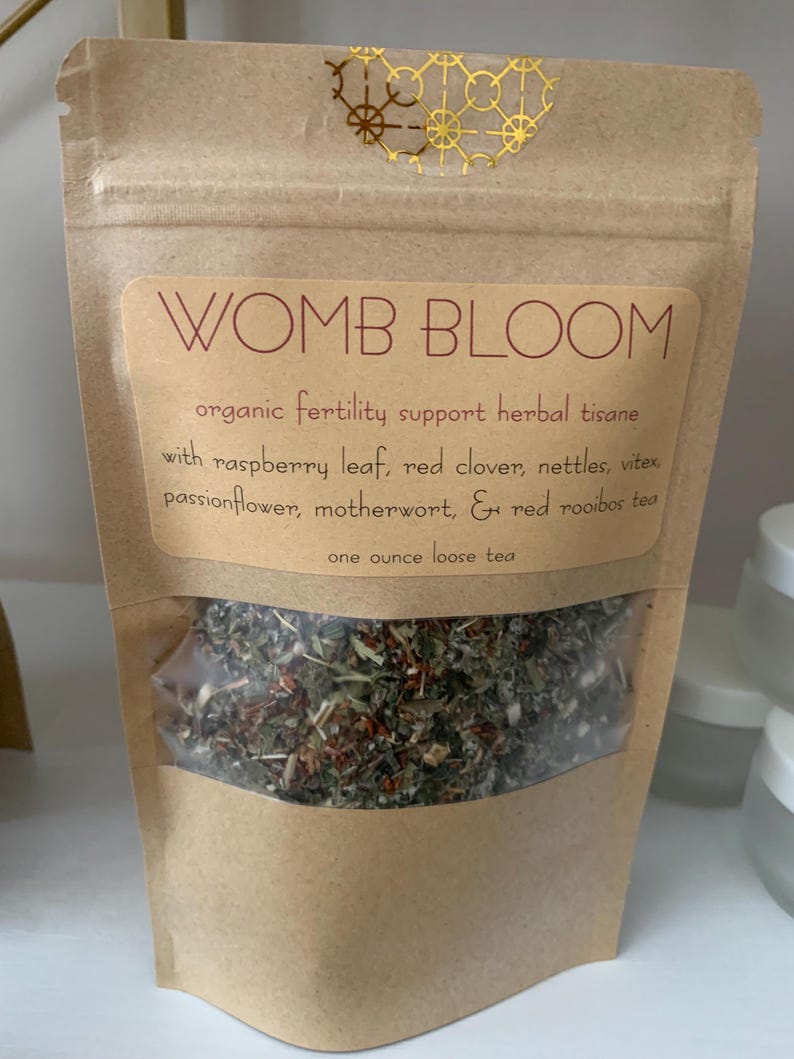 May include: A brown paper bag with a clear window displaying loose leaf tea. The label reads "WOMB BLOOM" and lists ingredients like raspberry leaf and red clover. The bag contains one ounce of tea.