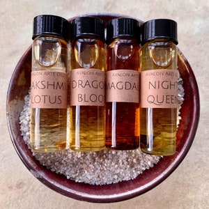 May include: Four small glass bottles with black caps, each containing a different colored liquid, are arranged in a maroon bowl filled with small, light-colored stones. The bottles are labeled "Lakshmi Lotus", "Dragon Blood", "Magdal", and "Night Queen".