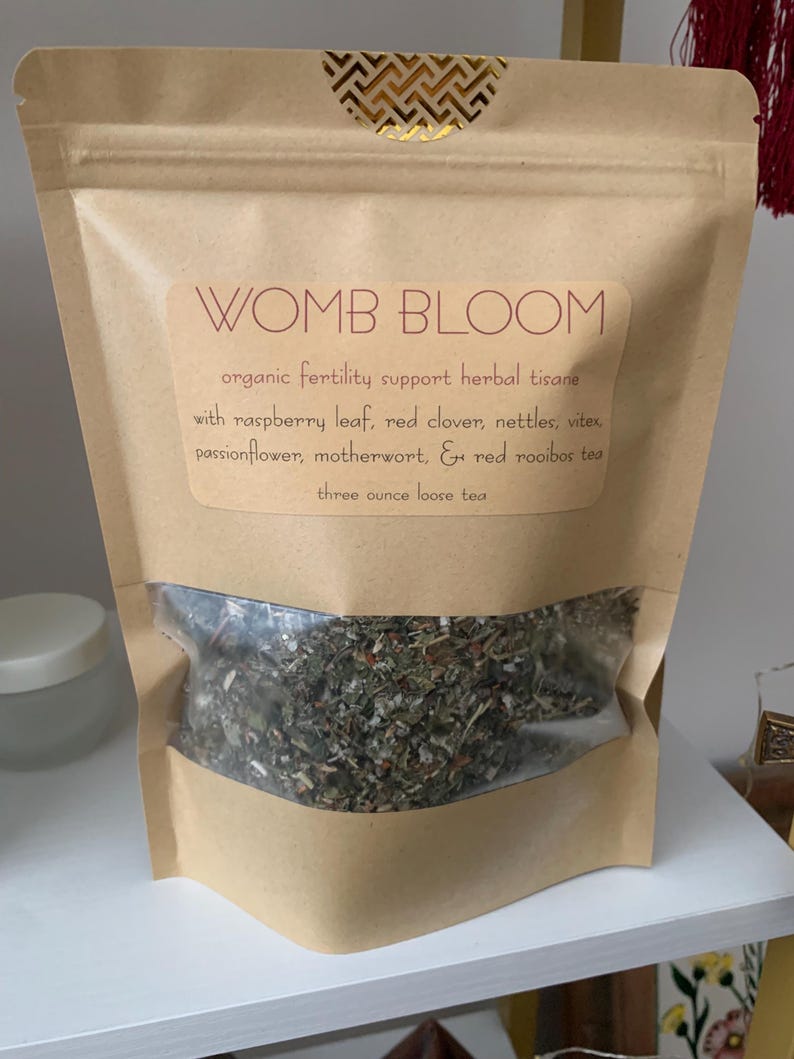 May include: A brown paper bag with a clear window displaying loose leaf tea. The label reads "WOMB BLOOM" and lists ingredients like raspberry leaf and red clover. The bag is sealed with a gold geometric design.