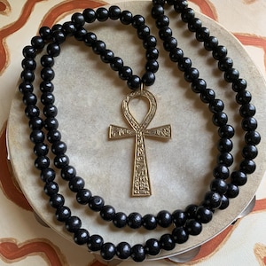 May include: A black beaded necklace with a gold-colored Ankh pendant. The necklace is made of round, dark beads and is draped over a light-colored surface with reddish-brown abstract designs. The Ankh pendant is detailed with etched patterns.