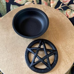 May include: A black, round bowl and a black pentagram-shaped coaster sit on a gold-colored surface. The bowl is empty, and the coaster features a five-pointed star design. The background includes a floral patterned fabric.