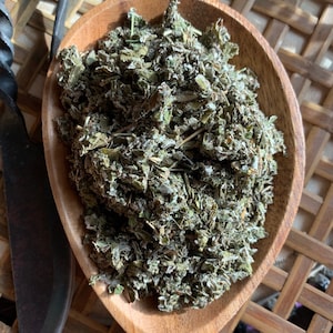 May include: A wooden scoop filled with dried green herbs. The scoop is oval-shaped and made of light-colored wood. A dark metal knife is visible on the left side of the image. The herbs have a textured appearance.