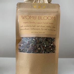 May include: A brown paper bag with a clear window, filled with loose herbal tea. The label reads "WOMB BLOOM" and lists ingredients like raspberry leaf and red clover. The bag contains one ounce of tea.