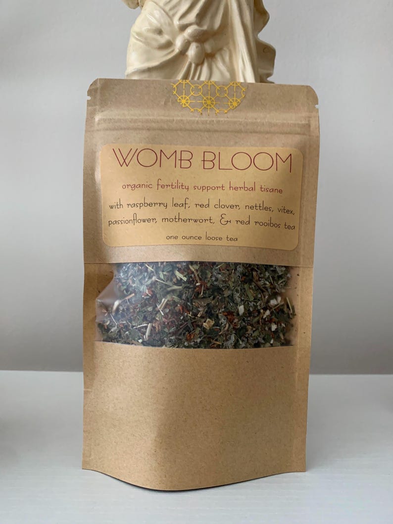 May include: A brown paper bag with a clear window, filled with loose herbal tea. The label reads "WOMB BLOOM" and lists ingredients like raspberry leaf and red clover. The bag contains one ounce of tea.