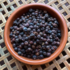 May include: A terracotta bowl filled with dark blue-purple juniper berries. The bowl sits on a woven bamboo mat. The berries are round and have a slightly wrinkled texture. The image is a close-up shot, highlighting the berries' color and texture.