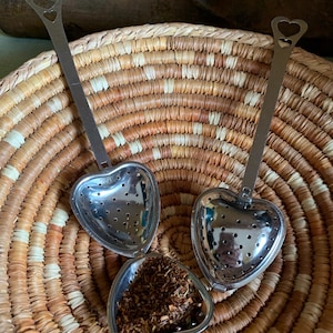 May include: Three silver heart-shaped tea infusers with handles. The infusers are sitting in a woven basket. One infuser is filled with loose leaf tea.