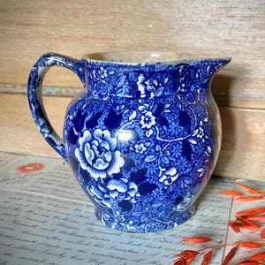 Puede incluir: Una jarra de cerámica con un patrón floral de transferencia en azul oscuro y blanco. El recipiente tiene un cuerpo redondeado, un asa curva en el lado izquierdo y un pequeño pico vertedor. El diseño intrincado cubre toda la superficie.
