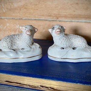 May include: Two white ceramic sheep figurines with textured wool, each resting on a white base with a thin gold trim. The sheep have painted facial features and are positioned in a reclining pose. One base shows a visible crack in the ceramic.